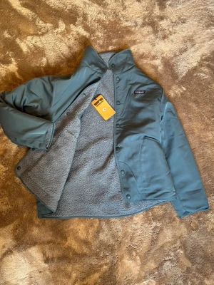 Grå teddyjacka från Patagonia - Säljer en grå teddyjacka från Patagonia med hög krage och tryckknappar. Jackan har en mjuk, fluffig utsida och slät insida, samt en bröstficka med dragkedja och två sidofickor. Perfekt för kyliga dagar och riktigt snygg till streetwear.