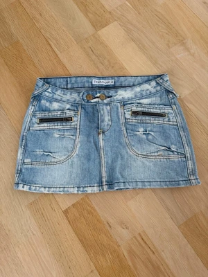 Ljusblå jeanskjol från Terranova - Säljer en ljusblå jeanskjol från Terranova med slitna detaljer och dubbla knappar fram. Kjolen har två framfickor med dragkedjor och bakfickor med broderade mönster. Klassisk denimlook med cool tvätt och kort passform.