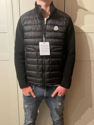 Moncler cardigan  - Säljer en exklusiv och mycket stilren jacka från Moncler. Jackan är en modern hybridmodell med vadderad front i lätt dunliknande material och stickade ärmar, vilket ger både värme och bra rörelsefrihet. Perfekt som övergångsjacka under vår/höst eller som ett extra lager under kyligare dagar.