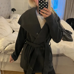 Grå omlottkappa från Orsay - Säljer en grå kappa från Orsay med bred krage och omlottmodell. Jackan har bälte i midjan och är helsvart med en stilren look. Perfekt för dig som gillar klassiska och snygga ytterplagg. Materialet känns mjukt och värmande. Står ingen storlek men skulle säga att den är i storlek s