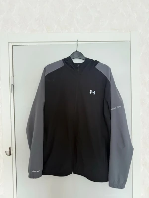 Svart/grå vindjacka från Under Armour - Säljer en svart och grå vindjacka från Under Armour med huva och dragkedja framtill. Jackan har logga på bröstet och text på ärmen. Tillverkad i lätt syntetmaterial, perfekt för blåsiga dagar. Sportig look och relaxed fit.