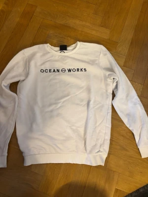 Vit sweatshirt från Ocean Works - Säljer en clean vit sweatshirt från Ocean Works i storlek M. Tröjan har rund halsringning, svart tryck med logga på bröstet och ribbade muddar vid ärmslut och nederkant. Perfekt för en enkel och stilren look.