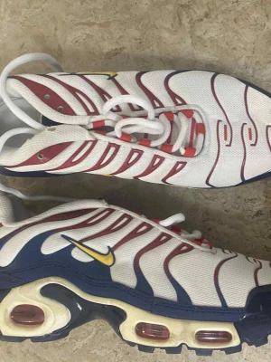 Nike Air Max Plus vita/röda/blå - Nike Air Max Plus sneakers i vitt mesh med röda och blå detaljer samt gula inslag på swoosh och Tn-logga. Skorna har synlig Air-enhet i sulan, vågiga röda linjer och vita snören. Snygg retrostil och riktigt sköna på foten.