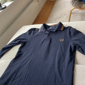 Mörkblå långärmad piké Fred Perry XS passar bra i S - Snygg mörkblå långärmad pikétröja från Fred Perry i bomull. Klassisk krage med bruna ränder och broderad logga i gult på bröstet. Perfekt för dig som gillar stilren och sportig vibe. Passar dig som vill ha en tidlös look med edge.