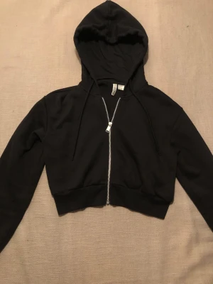Svart kort Hoodie från hm i storlek XS - Svart kort Hoodie med huva, dragkedja och snören från hm i storlek XS. Säljer denna tröjan för att den inte passar mig längre. Den är knappast använd. Du betalar frakten!