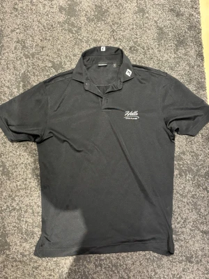 Svart pikétröja från FootJoy - Svart pikétröja från FootJoy med klassisk krage och korta ärmar. På bröstet finns en vit broderad text 'Hills Team Player'. Tröjan är tillverkad i ett lätt och syntetiskt material som andas, perfekt för sportiga aktiviteter.