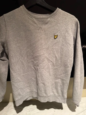 Grå sweatshirt från Lyle & Scott Junior - Klassisk grå sweatshirt från Lyle & Scott Junior med rund hals och broderad gul logga på bröstet. Tröjan har långa ärmar och ribbade muddar vid ärmslut och nederkant. Perfekt för en avslappnad och stilren look.