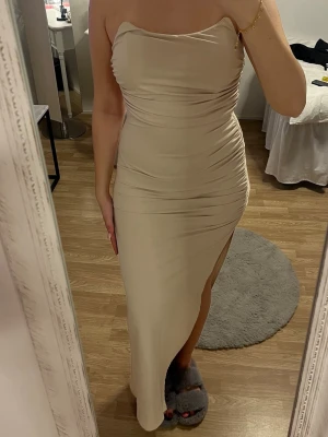 Beige axelbandslös långklänning  - Säljer en beige axelbandslös långklänning med tight passform och snygg slits på sidan. Klänningen har draperade detaljer längs sidorna. Aldrig använd och har prislapp kvar. Den är lång och går ner till mina fötter och jag är 170cm lång. 
