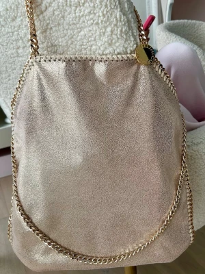 Guldig axelväska Stella McCartney - Säljer en snygg guldig axelväska från Stella McCartney med kedjedetaljer runt kanterna och axelrem. Väskan har en mjuk, glansig yta och är i den stora modellen. Perfekt för dig som vill ha en statementväska med lyxig känsla eller för att festa! 💖💖💖 skriv gärna vid intresse! 🩵🩵