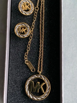 Smyckesset MK halsband & örhängen - Snyggt smyckesset från Michael Kors med halsband och matchande örhängen i guld- och silverfärg. Både halsbandet och örhängena har rund form med flätad silverdetalj runt MK-loggan. Kedjan är tunn och elegant med en liten bricka märkt K18 pläterad.