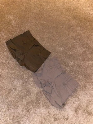 Två par shorts från H&M, strl 34 - Säljer två par shorts från H&M i storlek 34. Ena paret är olivgröna och det andra är beige, båda med fickor och knappdetaljer på sidorna. Perfekta för varma dagar och har en avslappnad passform.