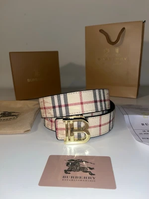 Rutig Burberry bälte med guldspänne - Säljer ett klassiskt Burberry bälte med ikoniskt rutigt mönster i beige, svart och rött. Bältet har ett stort guldigt B-spänne framtill och är gjort i skinn. Perfekt accessoar för att lyfta vilken outfit som helst.