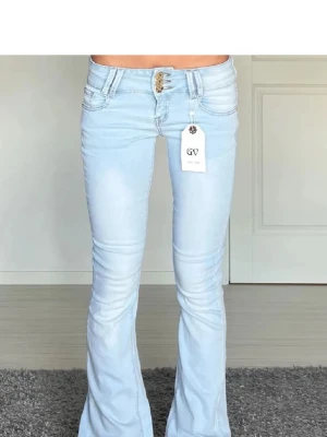 Ljusblå bootcut jeans från GV - Säljer ett par ljusblå jeans från GV med bootcut-snitt och låg midja. Jeansen har klassisk femficksdesign, snygga guldfärgade knappar och är tillverkade i mjukt denim-material. Perfekt för dig som gillar retrovibbar och vill sticka ut med en cool look.