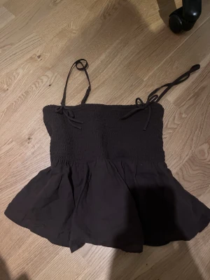 Brun babydoll från American Eagle - Säljer en brun linnetopp från American Eagle med smockad överdel och tunna axelband som knyts. Toppen har en volangig nederdel och är tillverkad i ett mjukt bomullstyg. Perfekt för varma dagar och enkel att styla med jeans eller kjol.