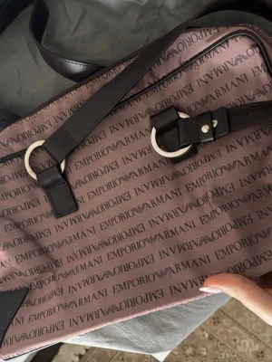 Emporio Armani väska i brunrosa - Säljer en snygg väska från Emporio Armani i brunrosa med logomönster över hela väskan. Väskan har svarta detaljer i skinn, silverfärgade metalldetaljer och dragkedja. Insidan är fodrad med svart tyg och har en innerficka med dragkedja.
