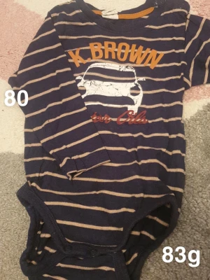 Randig långärmad body med biltryck - Mörkblå långärmad body med beige ränder och tryck av en bil. Praktiska knappar vid axeln och i grenen för enkel påklädning. Perfekt för dig som gillar coola och lekfulla plagg till de små.🌸Fint använt skick. Storlek 80