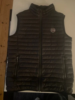 Svart dunväst från Colmar - Säljer en svart dunväst från Colmar Originals med quiltad design och hög krage. Västen har två fickor med dragkedja och Colmar-logga på bröstet. Insidan är ljusgrå och västen stängs med en rejäl dragkedja framtill. Perfekt för lager-på-lager och snygg till streetwear.