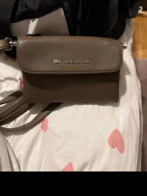 Brun axelväska från Michael Kors - Säljer en snygg brun axelväska från Michael Kors. Väskan har flera fack med dragkedja, silverfärgade detaljer och justerbar rem. Perfekt storlek för det viktigaste och stilren design med MK-logga både på dragkedjor och insida.