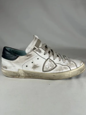 Vita Philippe model  - Säljer ett par vita sneakers från Philippe Model med grå snörning och svart hälparti. Skorna har en klassisk låg profil, rund tå och är tillverkade i skinn. Sidan pryds av märkets ikoniska sköldlogga. Perfekta för dig som gillar en avslappnad och trendig look.