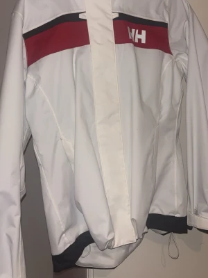 Vit vindjacka från Helly Hansen M - Säljer en vit vindjacka från Helly Hansen i storlek M. Jackan har en röd och svart detalj över bröstet med HH-logga, meshfoder på insidan och dragsko i nederkant. Stängs med dragkedja och har lång ärm. Perfekt för dig som vill ha en clean och sportig look. Använt några gånger sen mest legat i garderoben. Finns några märken men inget som syns om man inte kollar jätte noga. Skickar inom 1-2 dagar, ansvarar ej för postens slarv.