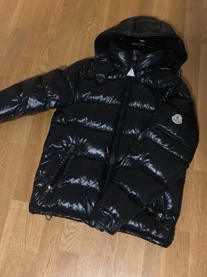 Svart pufferjacka Moncler - Säljer en svart, glansig pufferjacka från Moncler med huva och dragkedja framtill. Jackan har två snedställda fickor med dragkedja och Moncler-logga på ärmen. Perfekt för kalla dagar och riktigt snygg streetstil.