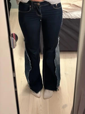 Mörkblå bootcut jeans med patchdetaljer - Unika mörkblå jeans med bootcut passform och stora ljusare denim-patchar med råa kanter längs benen och på sidan. Snygga kontrastsömmar och klassisk femficksdesign. Perfekta för dig som vill sticka ut med en edgy och trendig look.