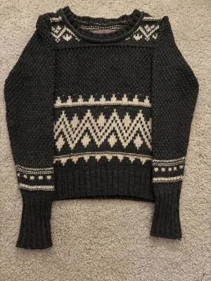 Mörkgrå stickad tröja med vitt mönster - Säljer en mörkgrå stickad tröja från Dr. Rehfeld med vita geometriska mönster över bröstet, axlarna och ärmarna. Tröjan har ribbade muddar och rund hals. Materialet är 100% akryl vilket gör den mjuk och varm. Perfekt för kalla dagar!