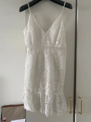 Vit spetsklänning från Love Triangle - Supersöt vit kortklänning från Love Triangle med tunna axelband och v-ringning. Klänningen har broderade spetsdetaljer och ett dekorativt mönster över hela tyget samt volang längst ner. Perfekt för sommarens alla tillfällen eller studenten! 