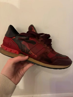 Valentino Rockrunner sneakers röd camo - Säljer ett par Valentino Rockrunner sneakers i vinrött och svart med camouflagemönster. Skorna har detaljer i mocka och skinn, samt nitar på hälen. Klassisk snörning och platt sula. Perfekta för dig som vill sticka ut med exklusiva och unika sneakers.