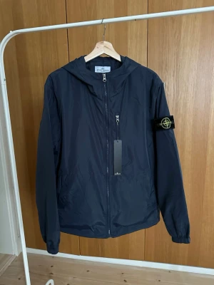 Mörkblå vindjacka från Stone Island - Snygg mörkblå vindjacka från Stone Island med huva och klassisk logotyp-patch på ärmen. Jackan har dragkedja framtill, en bröstficka med zip och elastiska muddar vid ärmslut. Perfekt för dig som vill ha en clean och stilren look.