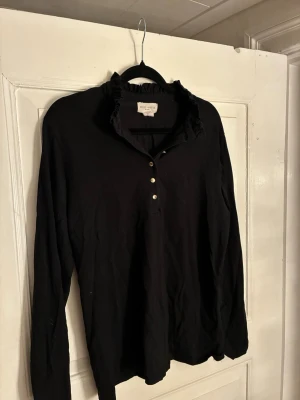 Svart tröja med volangkrage Holly & Whyte - Säljer en svart tröja från Holly & Whyte med lång ärm och volangdetaljer vid kragen. Tröjan har knappar framtill och är gjord i mjuk bomull. Perfekt till jeans eller kjol för en enkel men snygg look.