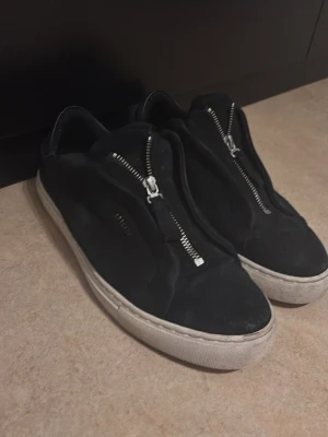 Svarta sneakers från Axel Arigato - Svarta sneakers från Axel Arigato i mocka med vit platt sula och silverfärgad dragkedja på ovansidan. Snygg minimalistisk design med rund tå och diskret logga på sidan. Perfekt för dig som gillar stilrena och moderna skor. Skick ligger på 7/10, kan även försöka putsa till dem lite till bara att höra av er vid fundering! 