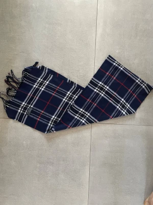 Burberry halsduk  - Tja! Säljer en klassisk rutig halsduk från Burberrys i mörkblått, vitt och rött. Halsduken är tillverkad i 100% lammull och har tvinnade fransar i ändarna. Perfekt accessoar för att lyfta din outfit med en tidlös och sjukt snygg outfit! Denna färg är väldigt eftertraktad och en av de snyggaste färgerna du kan ha! Skick 10/10 som helt ny! Halsduken är vintage och denna modell tillverkades senast 1999! Har du några frågor eller fundering är de bara att höra av sig! Mvh Premium fashion🤗🤩