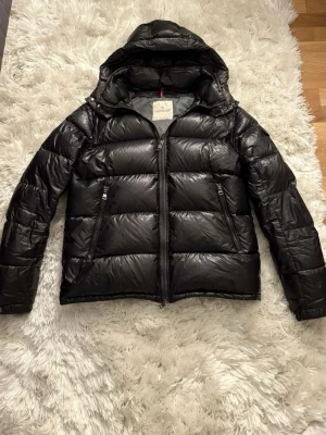 Svart Moncler pufferjacka med huva - Säljer en svart pufferjacka från Moncler med glansig finish och huva. Jackan har dragkedja framtill, två snedställda fickor med dragkedja och klassisk Moncler-etikett på insidan. Perfekt för kalla dagar och riktigt snygg streetstil.