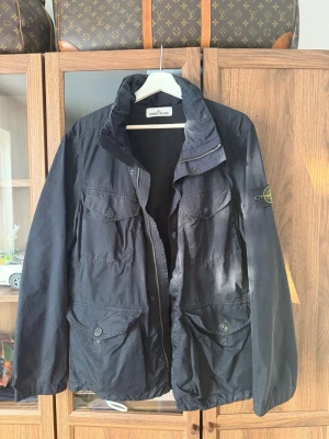 Mörkblå armyjacka från Stone Island - Säljer en mörkblå army/vindjacka från Stone Island med klassisk patch på ärmen. Jackan har fyra stora fickor framtill med knappar, hög krage och dragkedja. Perfekt till våren och hösten eller med en väst under. Super fint skick. Storlek L passas large och medium. Har uttagbar luva.