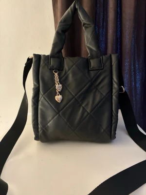 Svart quiltad axelväska med hjärtdetalj från H&M - Säljer en svart quiltad axelväska från H&M i syntetmaterial med både handtag och justerbar axelrem. Väskan har guldfärgade detaljer och en söt kedja med två hjärtberlocker framtill. Perfekt storlek för vardagsgrejer och ger en trendig look.