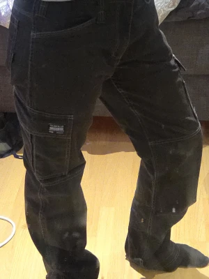 Svarta cargopants med fickor - Säljer ett par svarta cargopants med flera stora fickor på sidorna och vita kontrastsömmar. Byxorna har rak passform och är tillverkade i slitstarkt bomullstyg. Perfekta för dig som gillar streetwear och vill ha plats för allt i fickorna.