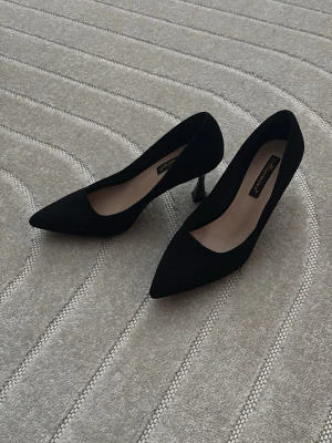 Svarta pumps med spetsig tå - Säljer ett par klassiska svarta pumps med spetsig tå och smal klack. Skorna är i mocka-look och har en elegant siluett som passar till många olika outfits. 