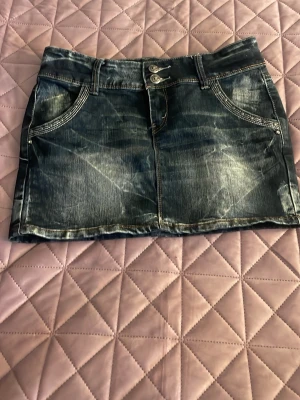 Mörk jeanskjol från Miss Grace - Snygg mörkblå jeanskjol från Miss Grace med slitningar, dubbla knappar fram och coola detaljer som dragkedjor och nitar på bakfickorna. Klassisk femficksmodell med bälteshällor och kontrastsömmar. Perfekt till sneakers eller boots. Midjemåttet är 38cm och är lågmidjade 