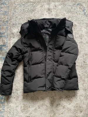 Svart Canada Goose jacka  - Säljer en svart Canada Goose jacka med huva och diskret logga på ärmen. Jackan har dragkedja och knappar framtill, samt flera praktiska fickor. Perfekt för kalla vinterdagar och riktigt snygg till alla outfits.