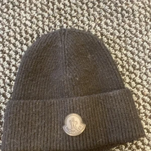 Svart Moncler mössa i ull - Svart ribbstickad mössa från Moncler med klassisk uppvikt kant och logotyp framtill. Tillverkad i mjuk ull som håller dig varm under kyliga dagar. Perfekt accessoar för att lyfta din vinterstil.