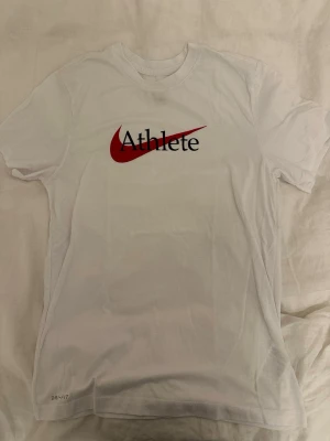 Vit Nike Dri-Fit Athlete t-shirt - Säljer en vit Nike t-shirt i Dri-Fit material med trycket 'Athlete' och en röd swoosh på bröstet. Klassisk rund hals och korta ärmar. Perfekt för dig som gillar sportig stil och vill ha något bekvämt och snyggt.
