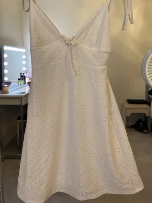 Mini vit känning  - Supersöt vit kort klänning med broderade hålmönster och smala axelband som knyts. V-ringad framtill med liten rosett och lätt utsvängd kjol. Perfekt för varma dagar och har en romantisk vibe. Materialet känns som bomull och är luftigt. Den är kort 