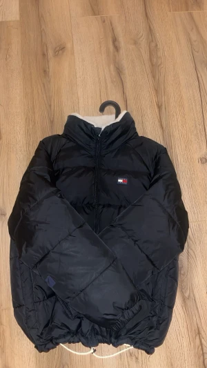 Svart pufferjacka från Tommy Jeans - Svart pufferjacka från Tommy Jeans med broderad logga på bröstet och vit teddyfoder i kragen. Jackan har dragkedja framtill, snörning i nederkant och quiltad design. Perfekt för kalla dagar och har en cool, oversized vibe. (HAR en liten defekt på undersidan armen inprincip onsylnig) PRISET ÄR INTE HUGGET I STEN!