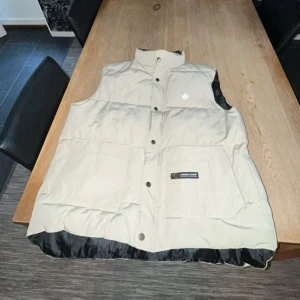 Beige dunväst från Canada Goose - Säljer en beige dunväst från Canada Goose med svart foder och broderad lönnlövdetalj. Västen har knappar framtill och praktiska fickor. Perfekt för kyliga dagar. Västen är oanvänd och i topp skick. Jag är 185 och den passar perfekt i längden. Är du 175 eller kortare kan de vara lite stor.