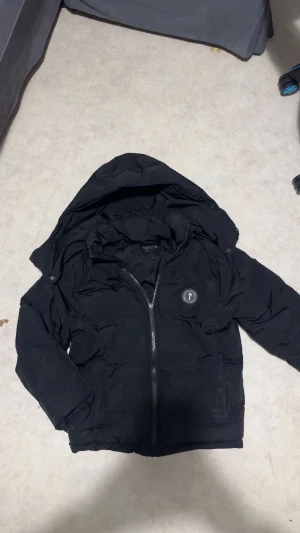 Svart pufferjacka från Trapstar - Svart pufferjacka från Trapstar med stor huva och broderad logga på ryggen. Jackan har dragkedja framtill, två sidofickor och rund patch med Trapstar-emblem på bröstet. Materialet är syntet och jackan har en loose passform, perfekt för kalla dagar. OBS! Den saknar dragkedja dragare