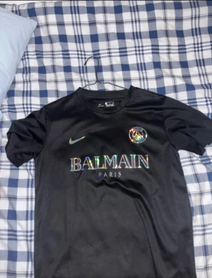Svart PSG x Balmain fotbollströja Nike - Svart fotbollströja från Nike med PSG och Balmain Paris-logga i holografiskt tryck på bröstet. Klassisk rund hals och korta ärmar. Tillverkad i 100% polyester för en sportig känsla. Perfekt för dig som gillar exklusiva samarbeten och snygg design.
