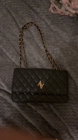 Svart quiltad axelväska från AV - Snygg svart axelväska från AV med quiltat mönster och guldfärgad kedja. Väskan har ett elegant AV-emblem framtill och praktiskt innerfack. Perfekt storlek för det viktigaste och ger en lyxig känsla till din outfit.