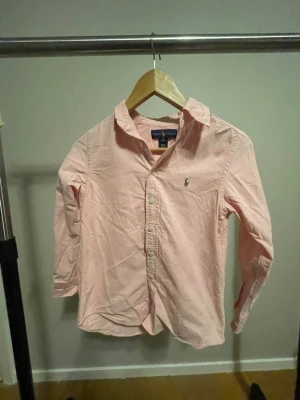 Rosa skjorta från Ralph Lauren - Snygg ljusrosa skjorta från Ralph Lauren med klassisk krage och broderad logga på bröstet. Skjortan har långa ärmar, knäppning framtill och är tillverkad i mjuk bomull. Perfekt för dig som gillar stilrena och tidlösa plagg.