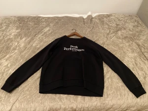 Svart Peak Performance sweatshirt - Svart sweatshirt från Peak Performance med vit logotyp på bröstet. Tröjan har rund hals, långa ärmar och ribbade muddar vid ärmslut och nederkant. Perfekt för en chill och sportig look, passar dig som gillar stilren design.